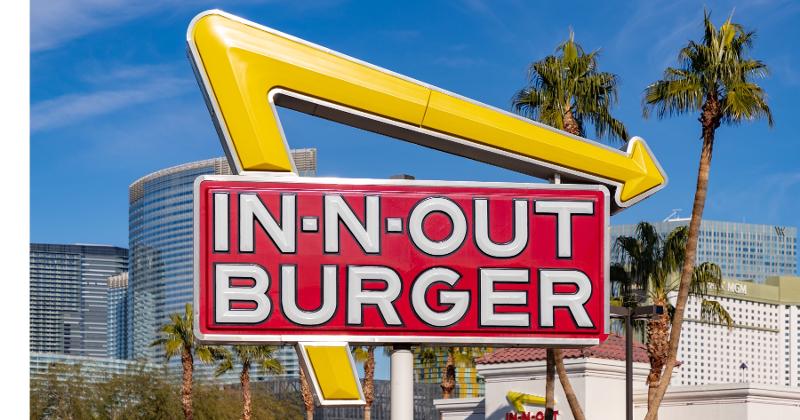 A daytime image of an In-N-Out Burger logo with palm trees and Las Vegas strip buildings in the background.